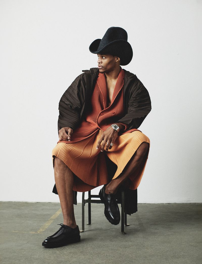 Russell Westbrook wearing a wide-brimmed black hat, pleated orange cardigan and shorts, and dark shoes while seated.