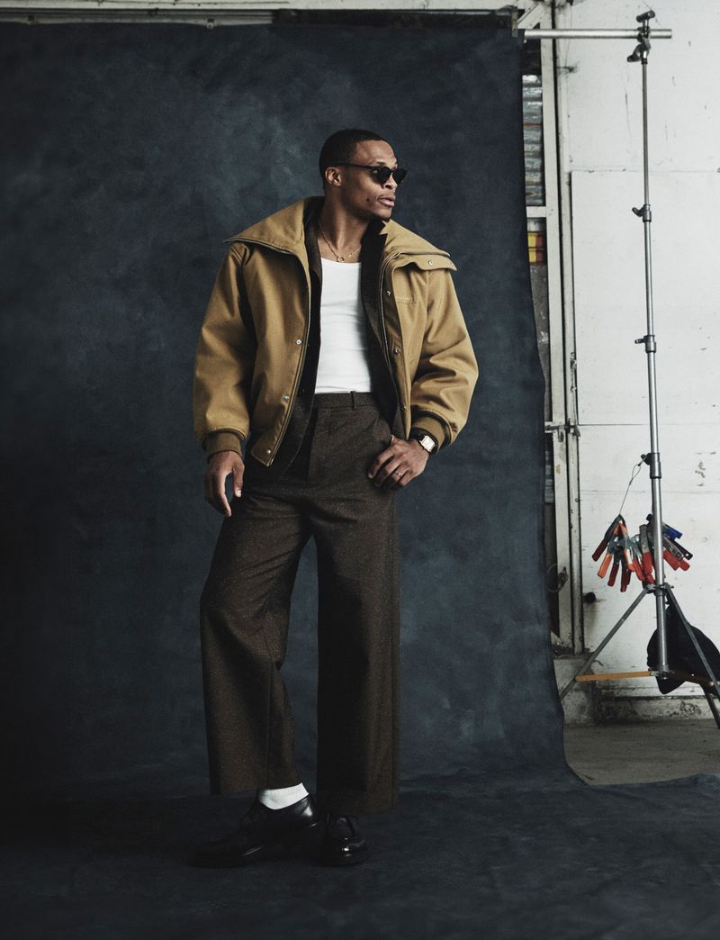 Russell Westbrook wearing a tan jacket, dark trousers, and sunglasses standing in front of a dark textured studio backdrop.