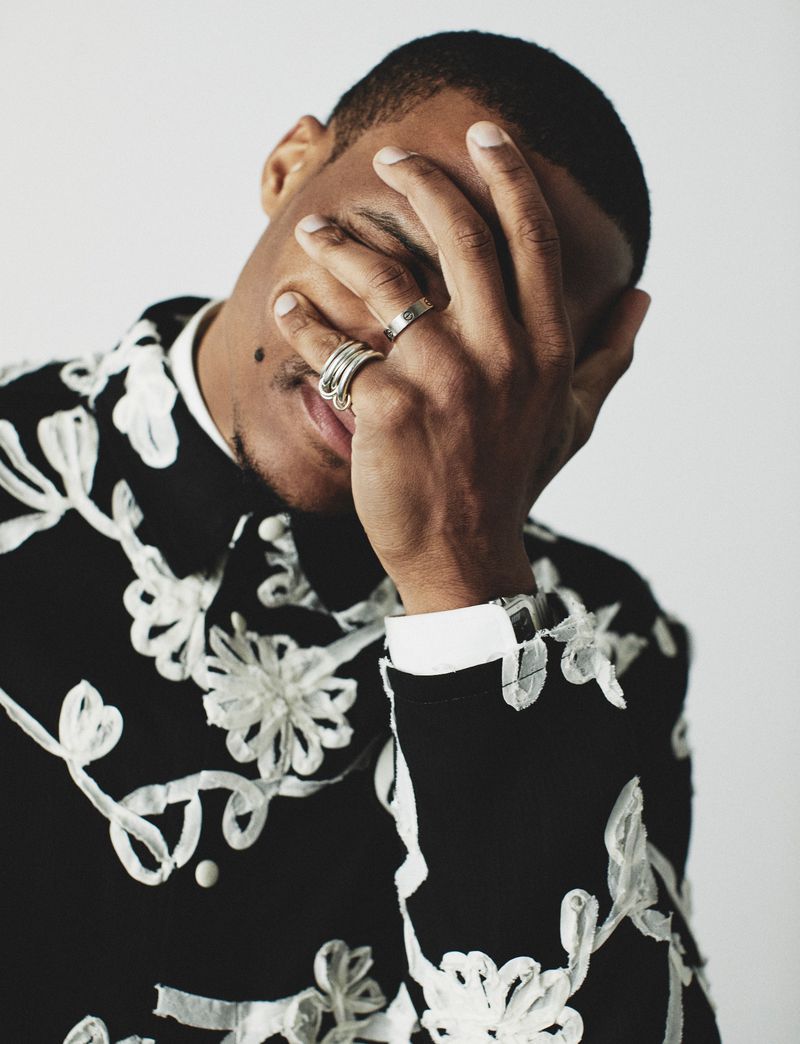 Russell Westbrook covering his face with his hand wearing silver rings and an embroidered black and white shirt against a white backdrop.