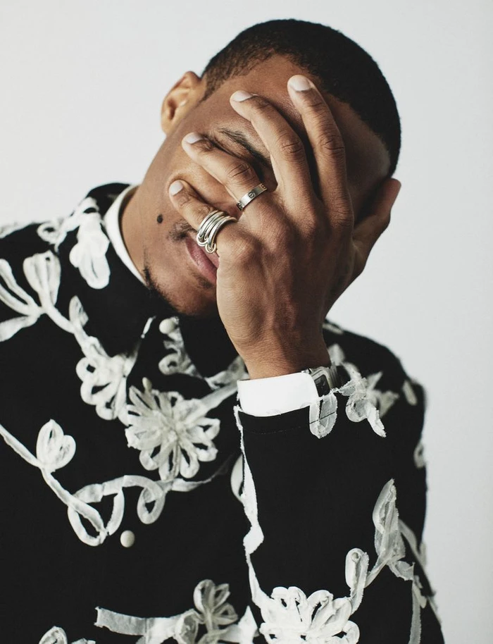 Russell Westbrook covering his face with his hand wearing silver rings and an embroidered black and white shirt against a white backdrop.