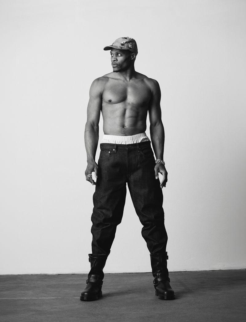Full-body black and white portrait of athlete Russell Westbrook standing shirtless in dark trousers and a cap against a studio backdrop.