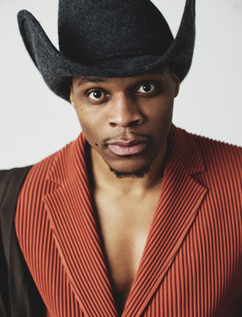 Russell Westbrook wearing a black cowboy hat and a rust pleated jacket against a white background.
