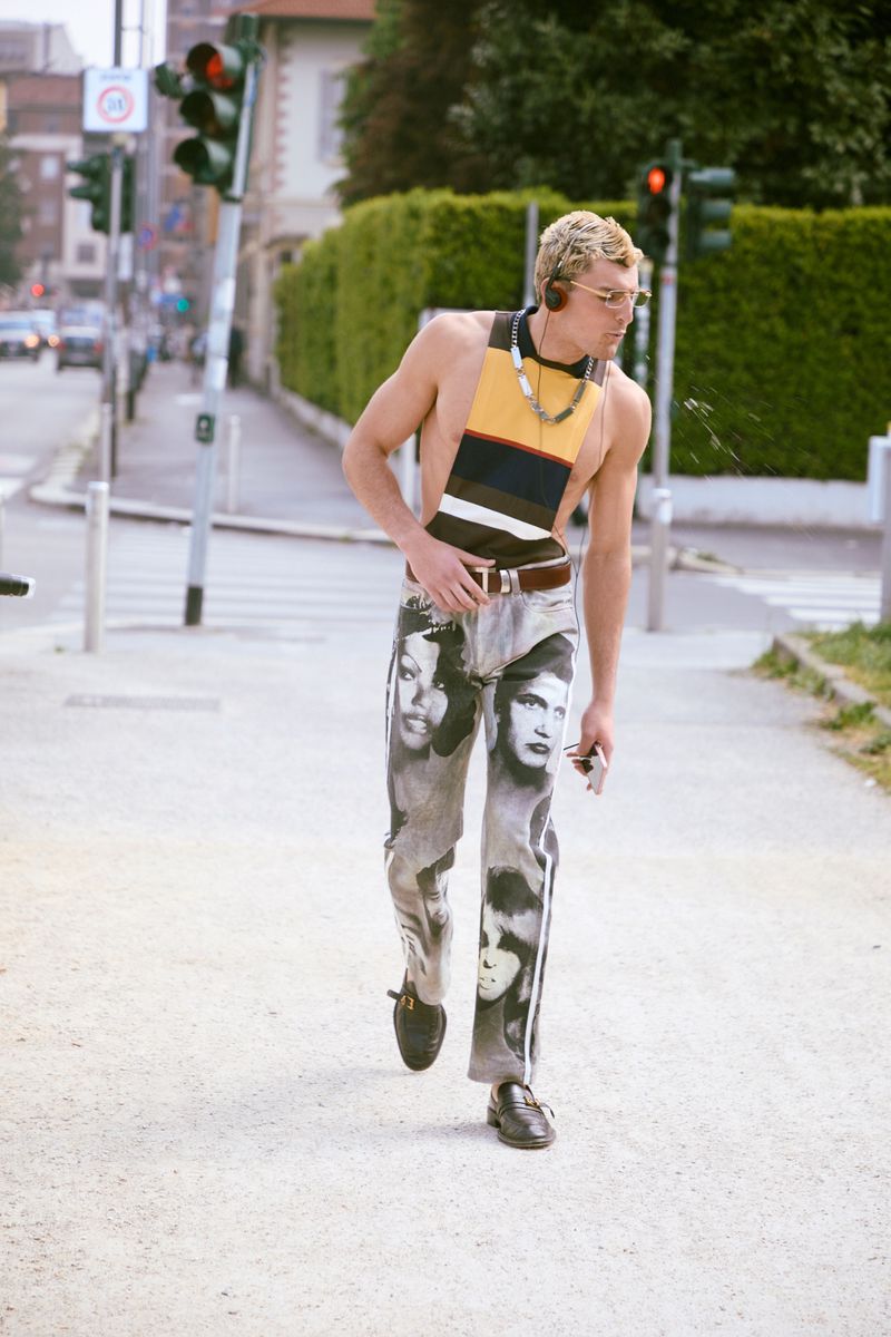 A man walks along a street wearing printed portrait pants and a striped halter top while spitting water.