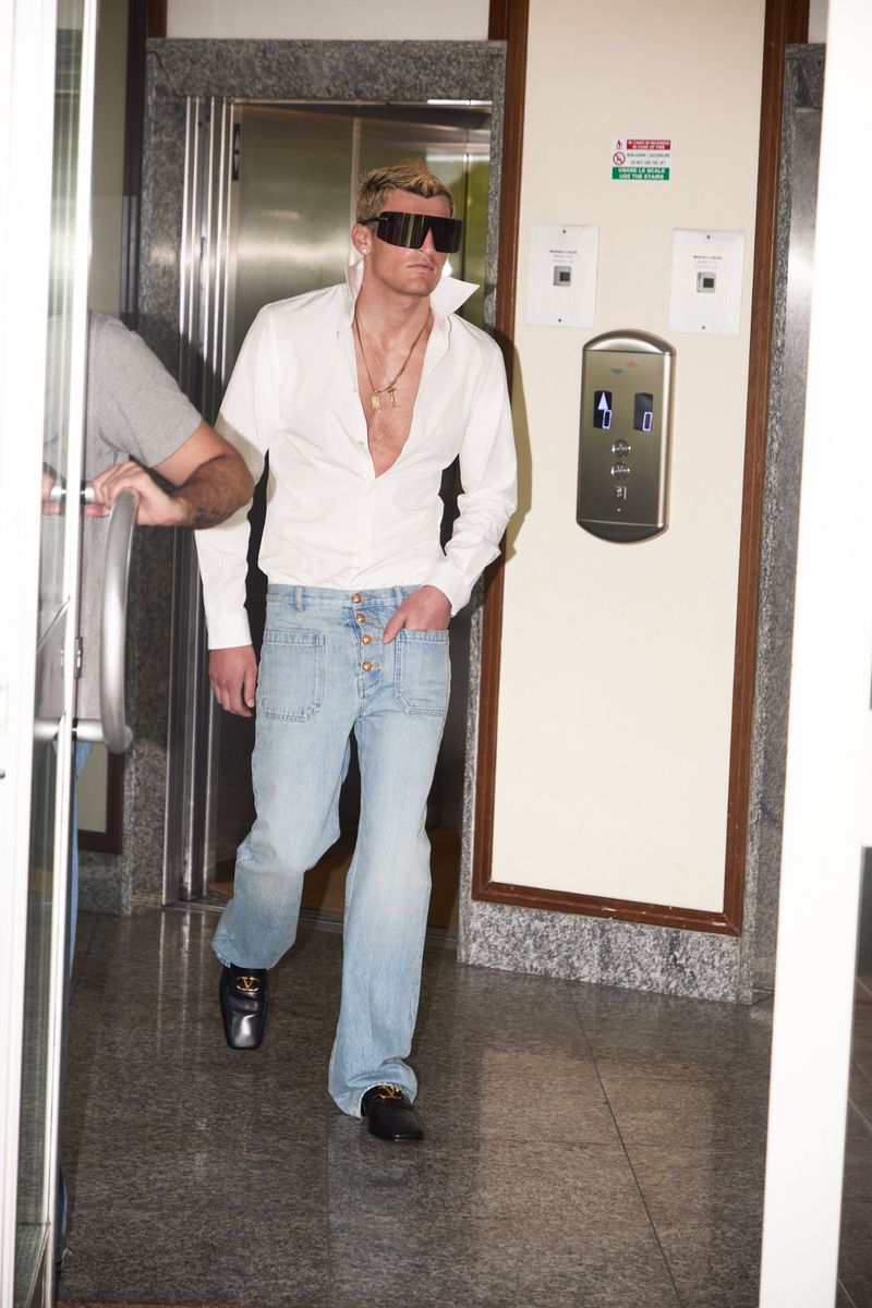 Man in oversized black sunglasses, an unbuttoned white collared shirt, and light blue denim jeans walking out of an elevator lobby.