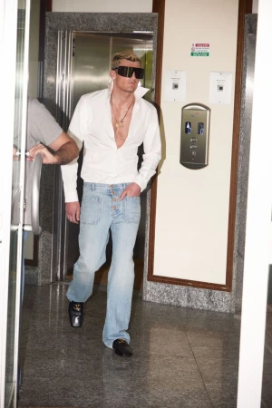 Man in oversized black sunglasses, an unbuttoned white collared shirt, and light blue denim jeans walking out of an elevator lobby.