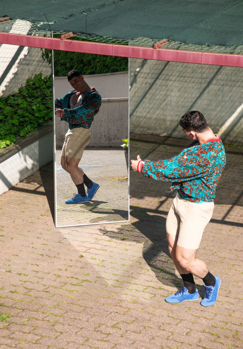 Man in a teal patterned jacket and beige shorts posing in front of a standing mirror outdoors on a paved courtyard.