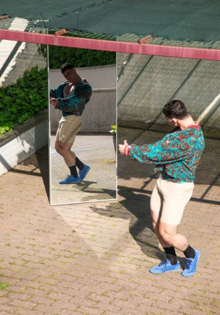 Man in a teal patterned jacket and beige shorts posing in front of a standing mirror outdoors on a paved courtyard.