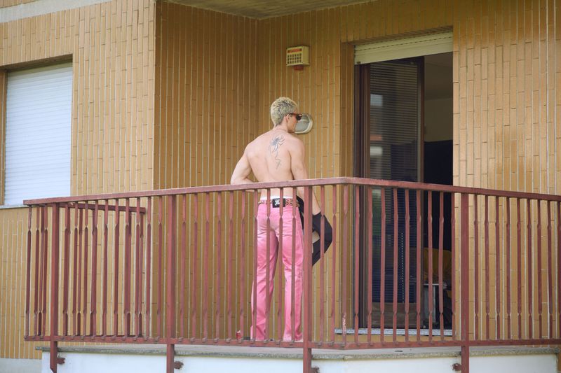 Shirtless man with blonde hair and back tattoo wearing pink trousers standing on an apartment balcony.