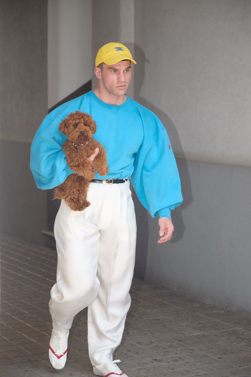 A man holding a small brown dog while walking in a bright blue sweatshirt, white pleated trousers, and a yellow cap.