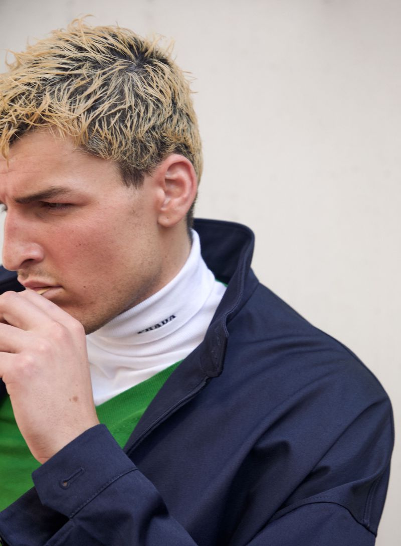 Profile portrait of a man with short blonde hair wearing a navy jacket, green top, and white turtleneck.