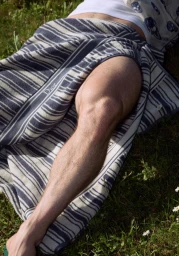 Close-up of a bare muscular leg resting on green grass, draped in a navy and off-white striped Dior towel under bright natural sunlight.