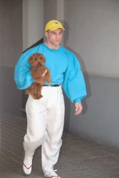 A man holding a small brown dog while walking in a bright blue sweatshirt, white pleated trousers, and a yellow cap.