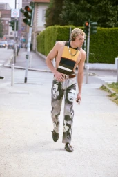 A man walks along a street wearing printed portrait pants and a striped halter top while spitting water.