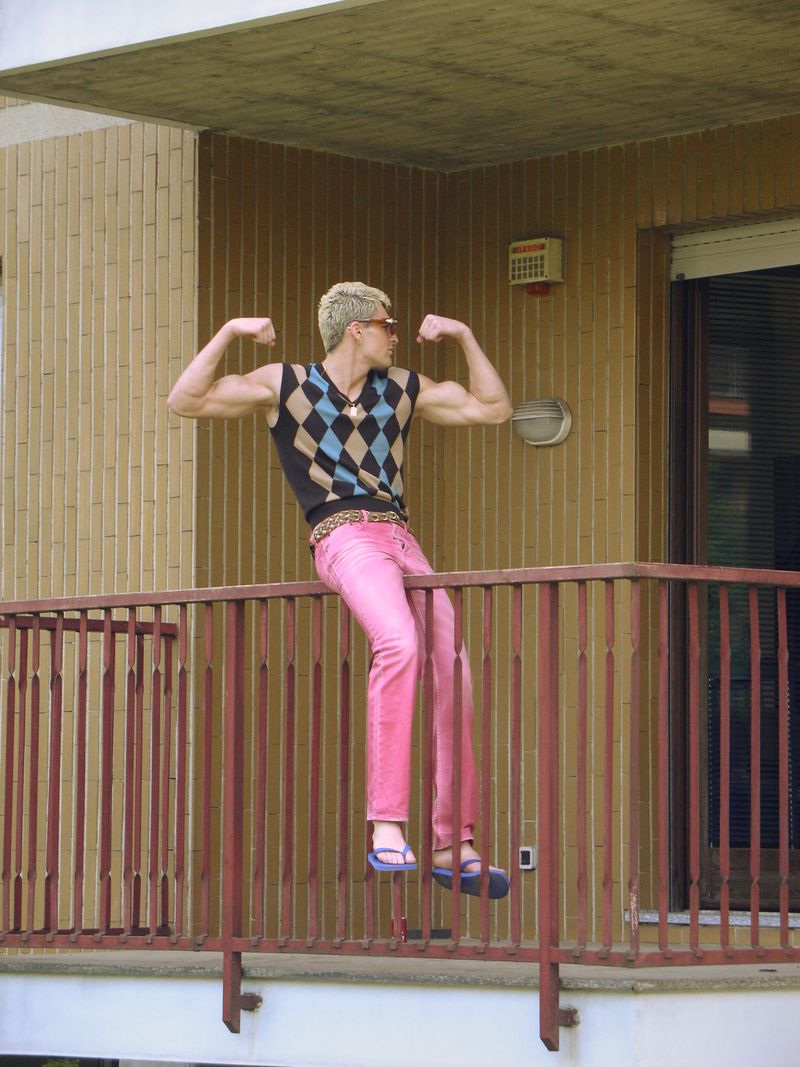 A man with blonde hair sitting on a balcony railing flexing his biceps in a sleeveless argyle sweater vest and pink trousers.