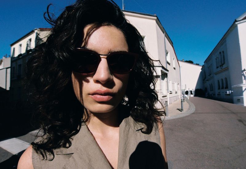 Close-up portrait of model Baptiste Vivo wearing dark sunglasses and a sleeveless beige top on a sunlit street.