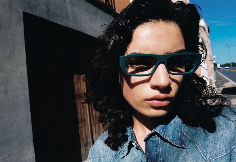 Close-up portrait of a model with dark curly hair wearing teal rectangular sunglasses and a blue denim shirt under direct sunlight.