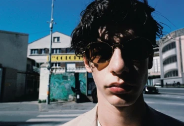 Close-up portrait of a young man wearing round sunglasses and a jacket outdoors on a city street.