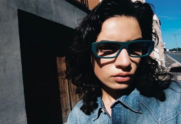 Close-up portrait of a model with dark curly hair wearing teal rectangular sunglasses and a blue denim shirt under direct sunlight.