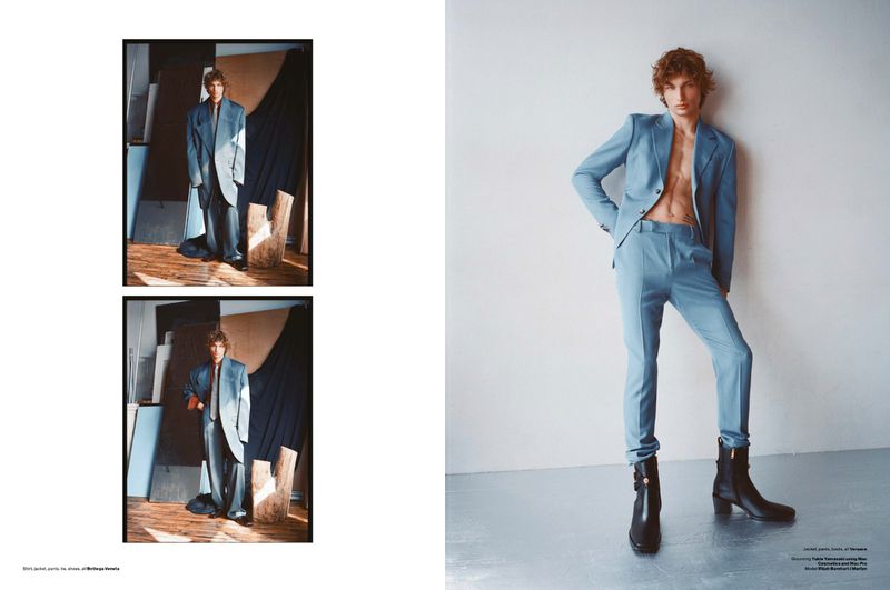 Editorial fashion shot of a young adult male model wearing a light blue suit and black boots against a minimalist backdrop.