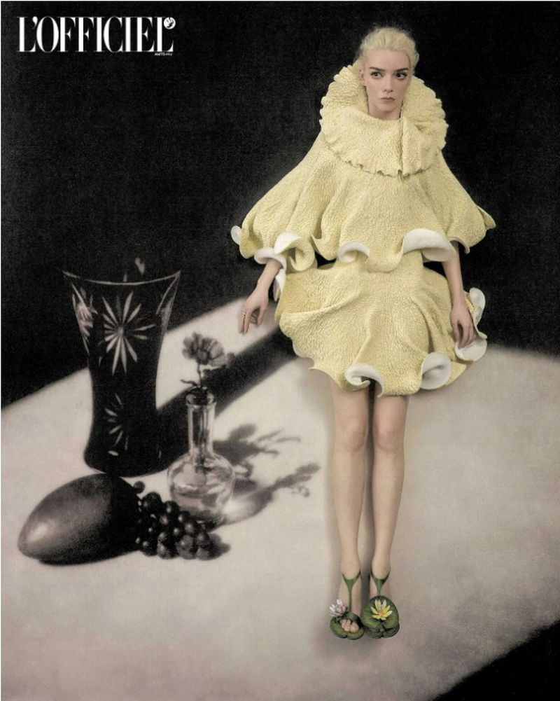 Anya Taylor-Joy wearing a sculptural yellow ruffle mini dress and flower heels next to a still life arrangement against a dark backdrop.