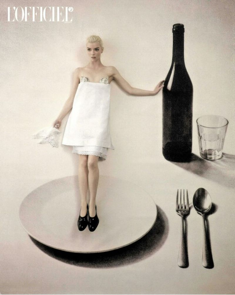 Anya Taylor-Joy in a white napkin dress standing on an oversized plate next to a giant wine bottle, glass, fork, and spoon.