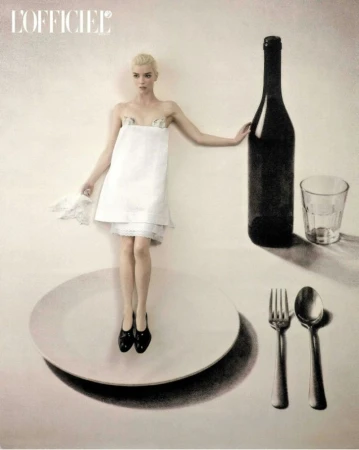Anya Taylor-Joy in a white napkin dress standing on an oversized plate next to a giant wine bottle, glass, fork, and spoon.