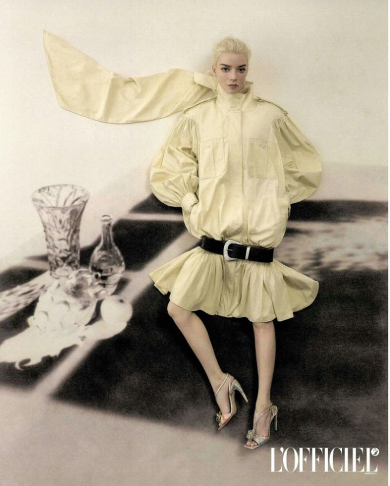 Anya Taylor-Joy posing in a voluminous pale yellow coat dress with a wide black belt against a graphic set.
