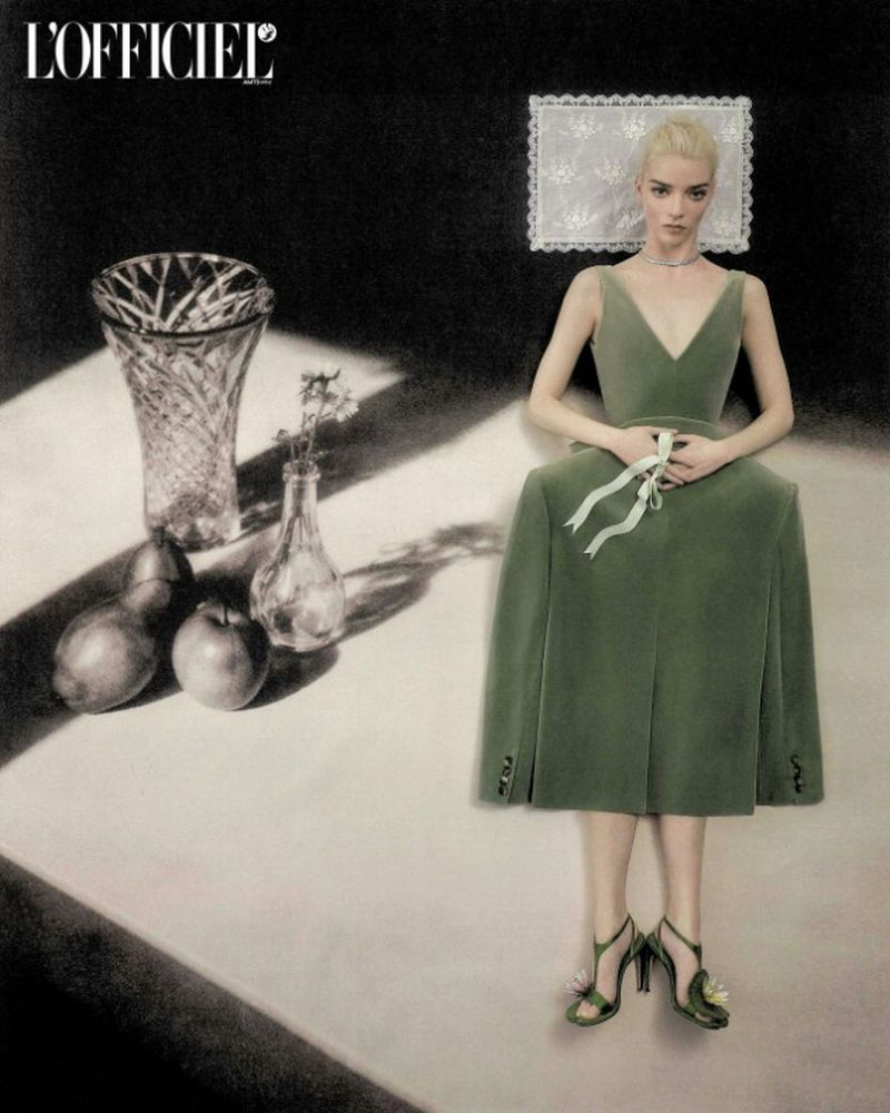 Anya Taylor-Joy poses in a green velvet dress beside a still life arrangement of crystal glassware and fruit.