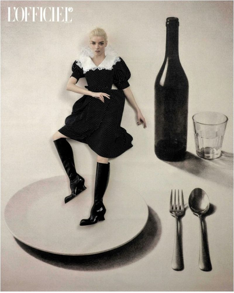 Anya Taylor-Joy in a black polka-dot dress and boots posing atop a large dinner plate beside giant tableware and a wine bottle.