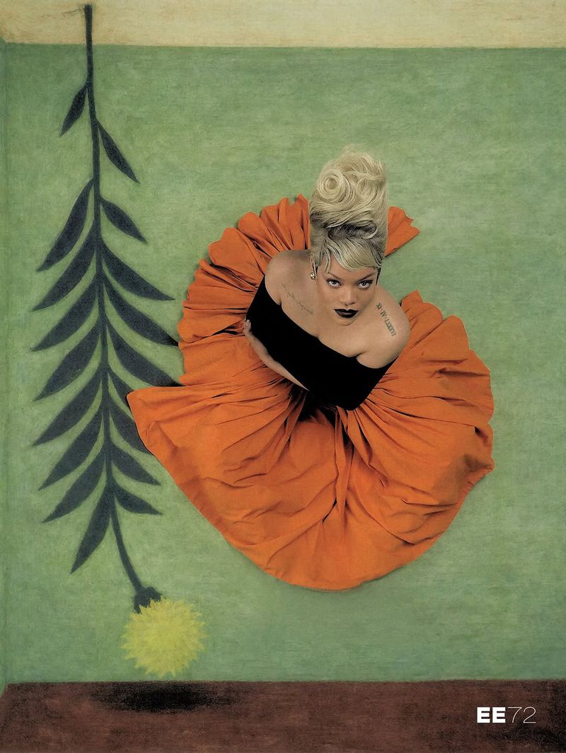 High-angle view of Rihanna looking upward, dressed in a voluminous orange skirt and black off-the-shoulder top beside a painted floral stem.