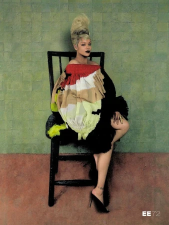 Rihanna seated on a black wooden chair wearing a colorful ruffled off-the-shoulder dress with a tall blonde updo against a green backdrop.
