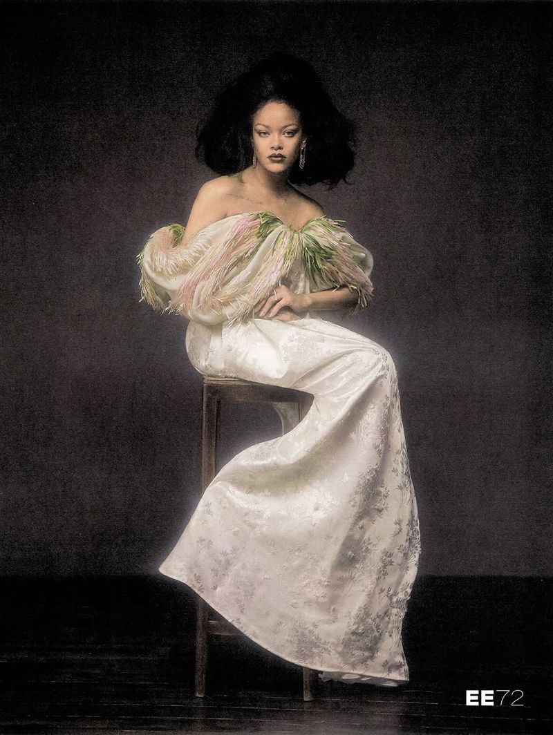 Rihanna seated on a wooden stool wearing a draped ivory off-the-shoulder gown with fringed neckline against a dark textured backdrop.