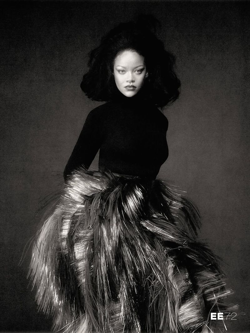 Rihanna posed in a fitted black turtleneck and a voluminous metallic fringe skirt against a dark textured backdrop.