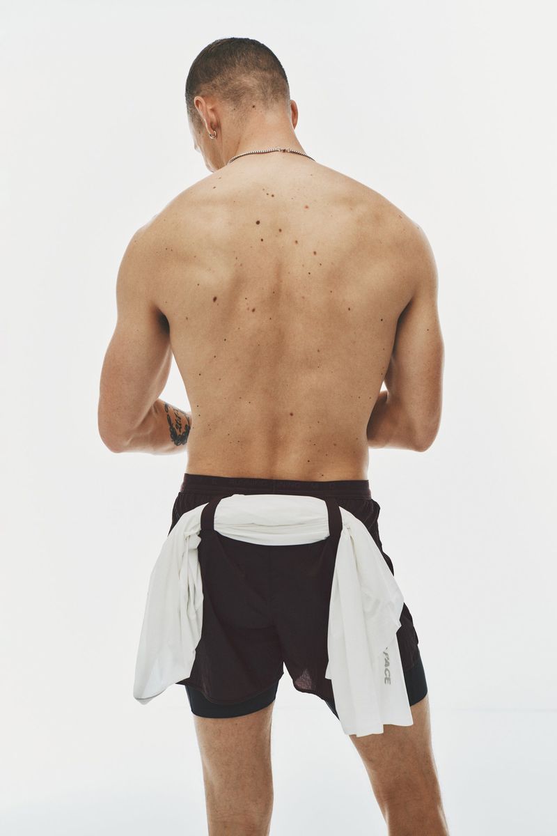 Rear view of a shirtless man wearing black athletic shorts with a white shirt tied around his waist against a white studio background.