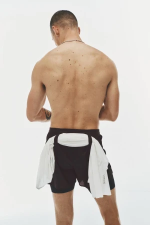 Rear view of a shirtless man wearing black athletic shorts with a white shirt tied around his waist against a white studio background.