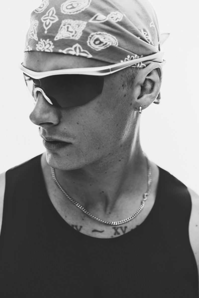 Close-up black and white portrait of a man wearing a patterned bandana, wraparound sunglasses, and a tank top against a plain background.