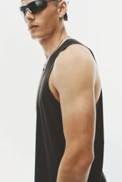 Male model wearing a black sleeveless tank top and sporty wrap-around sunglasses against a bright white background.