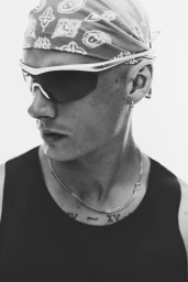Close-up black and white portrait of a man wearing a patterned bandana, wraparound sunglasses, and a tank top against a plain background.