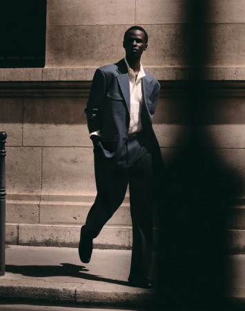 Malick Bodian walks along a sunlit city pavement wearing a relaxed dark grey suit and unbuttoned off-white collared shirt.