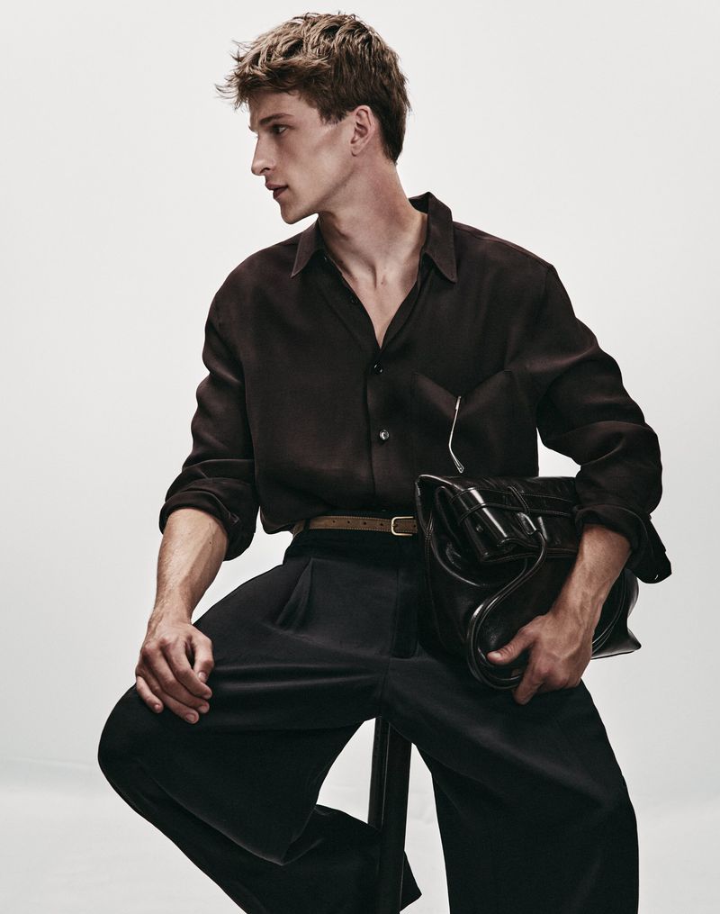 Model Vasko Luyckx seated in a dark brown collared shirt and black trousers holding a leather bag against a studio background.
