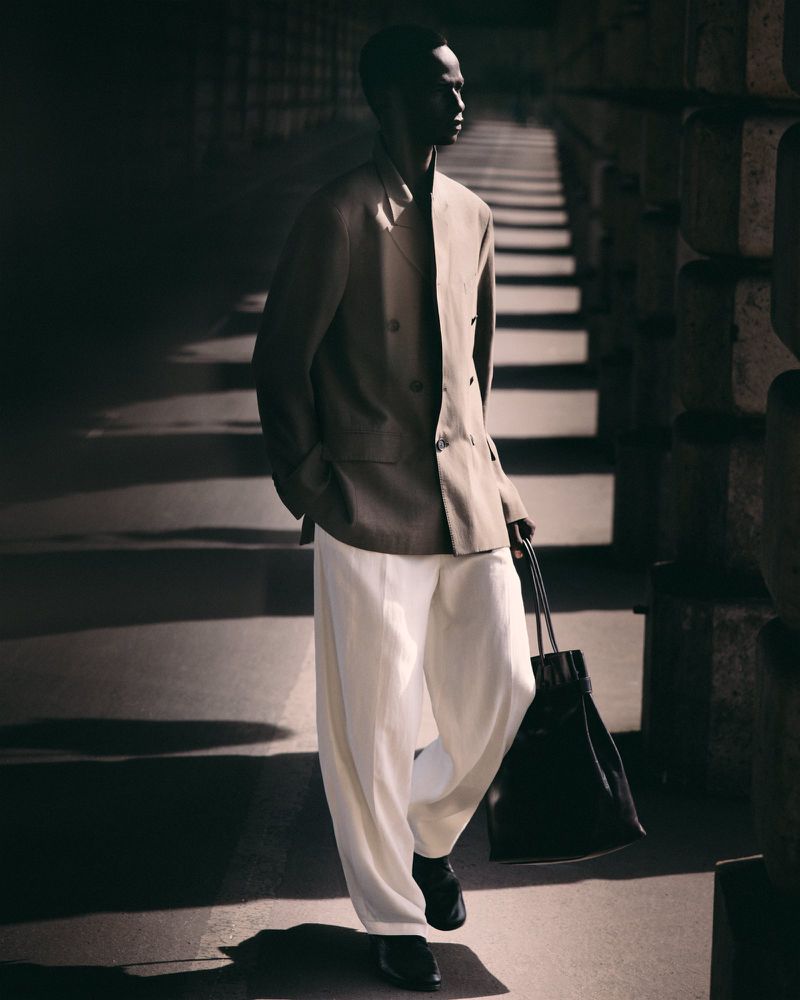 Black model wearing a double-breasted beige blazer and white trousers walking in shadow while holding a black tote bag.