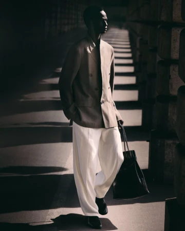 Black model wearing a double-breasted beige blazer and white trousers walking in shadow while holding a black tote bag.