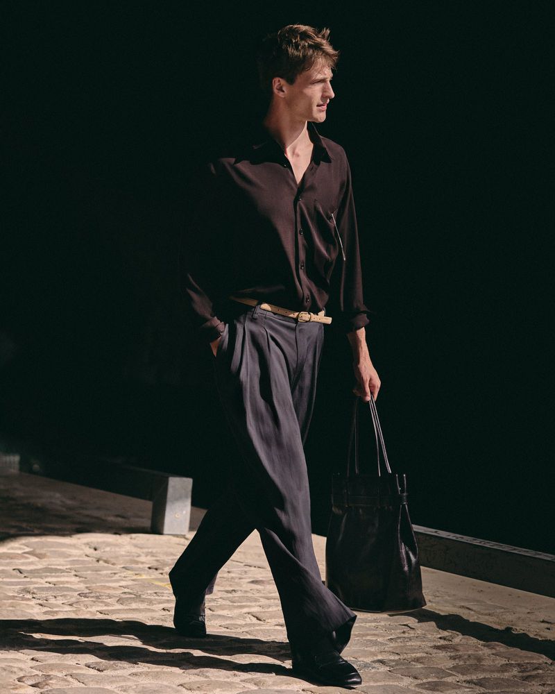 Model Vasko Luyckx walks along sunlit cobblestones wearing a dark button-down shirt, pleated trousers, and a black leather tote bag.