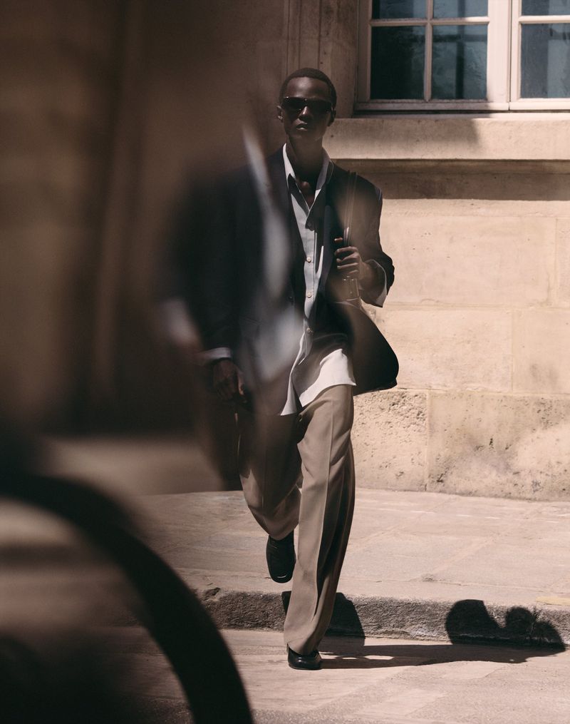 Malick Bodian walks outdoors wearing an oversized blazer, white shirt, wide-leg beige trousers, and dark sunglasses.