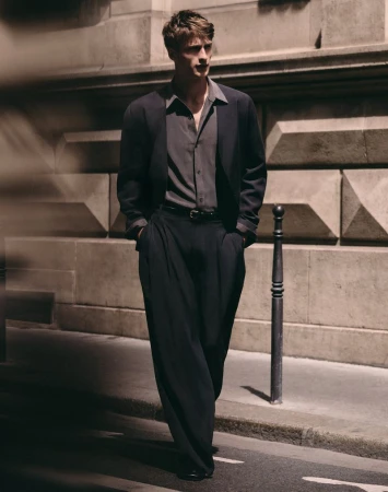 Model Vasko Luyckx walking down a city sidewalk wearing a dark tailored blazer, unbuttoned collared shirt, and wide-leg trousers.