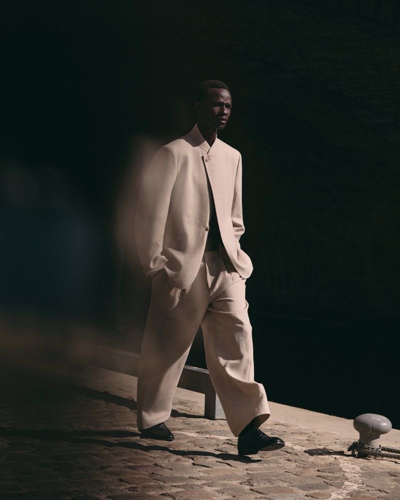 Model Malick Bodian walking on cobblestone in a tailored loose beige suit under directional sunlight with a dark backdrop.