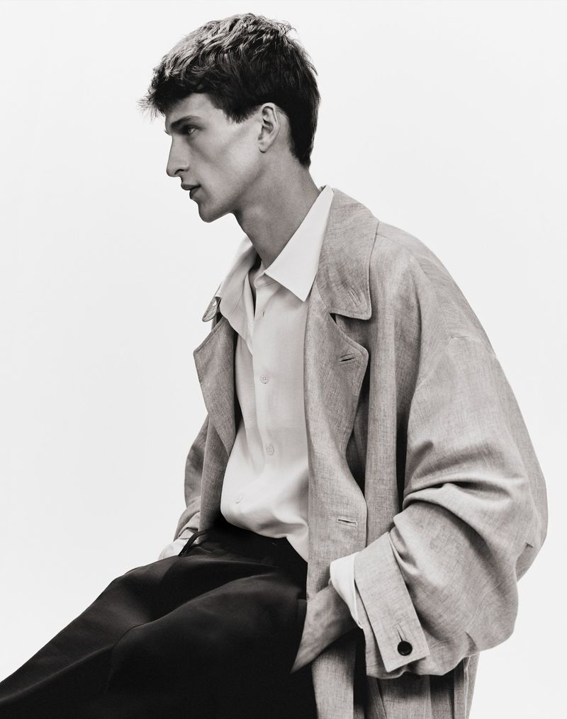 Male model in profile wearing an oversized trench coat, white collared shirt, and dark trousers against a white seamless studio backdrop.