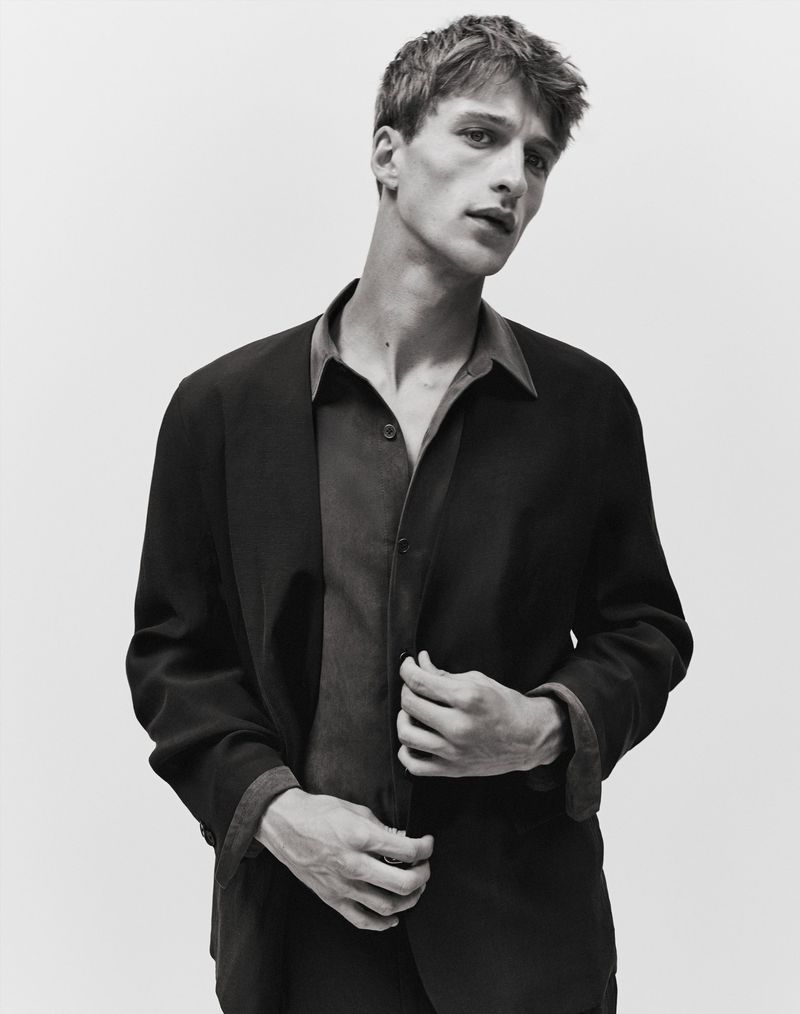 Monochrome portrait of a young man wearing a dark blazer over a collared shirt against a light seamless background.