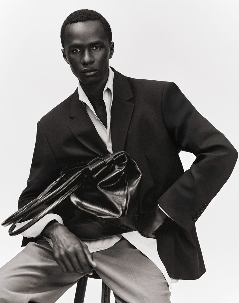 Model Malick Bodian seated in a black blazer holding a black leather tote bag against a white studio background.