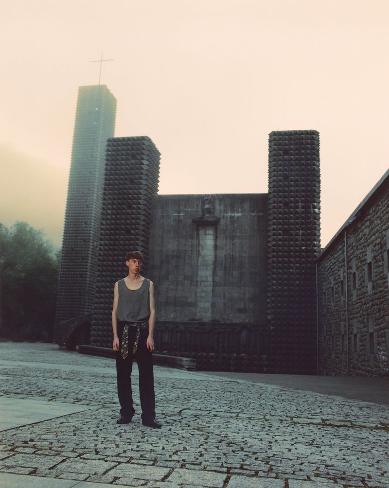Model wearing a grey tank top and dark trousers standing in a cobblestone courtyard before a brutalist stone church in misty weather.